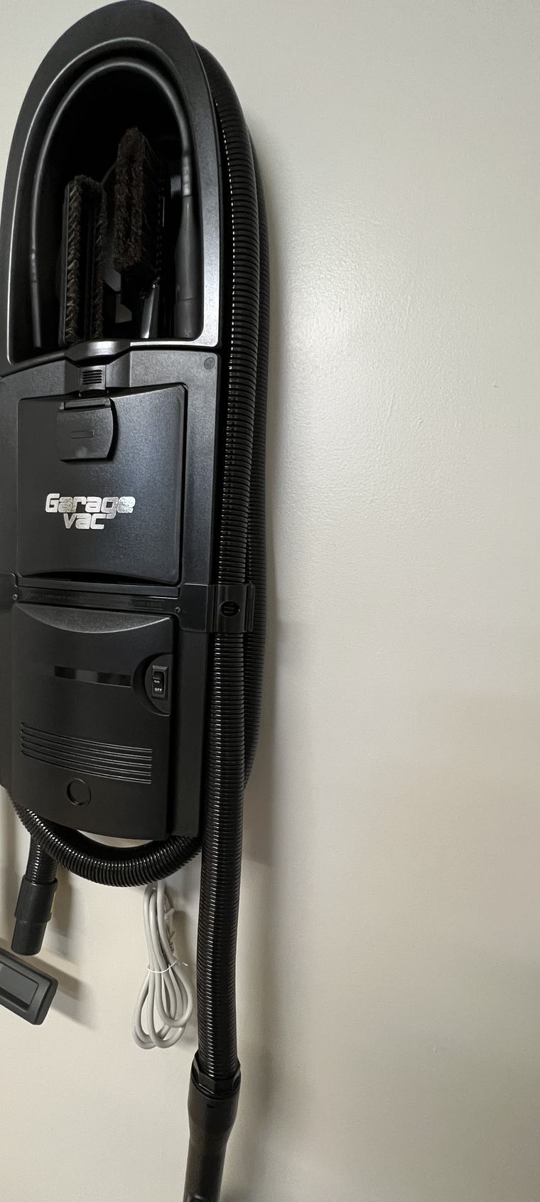 A GarageVac in ebony mounted on a garage wall, hose wrapped around the body and tools stored in the caddy at the top.