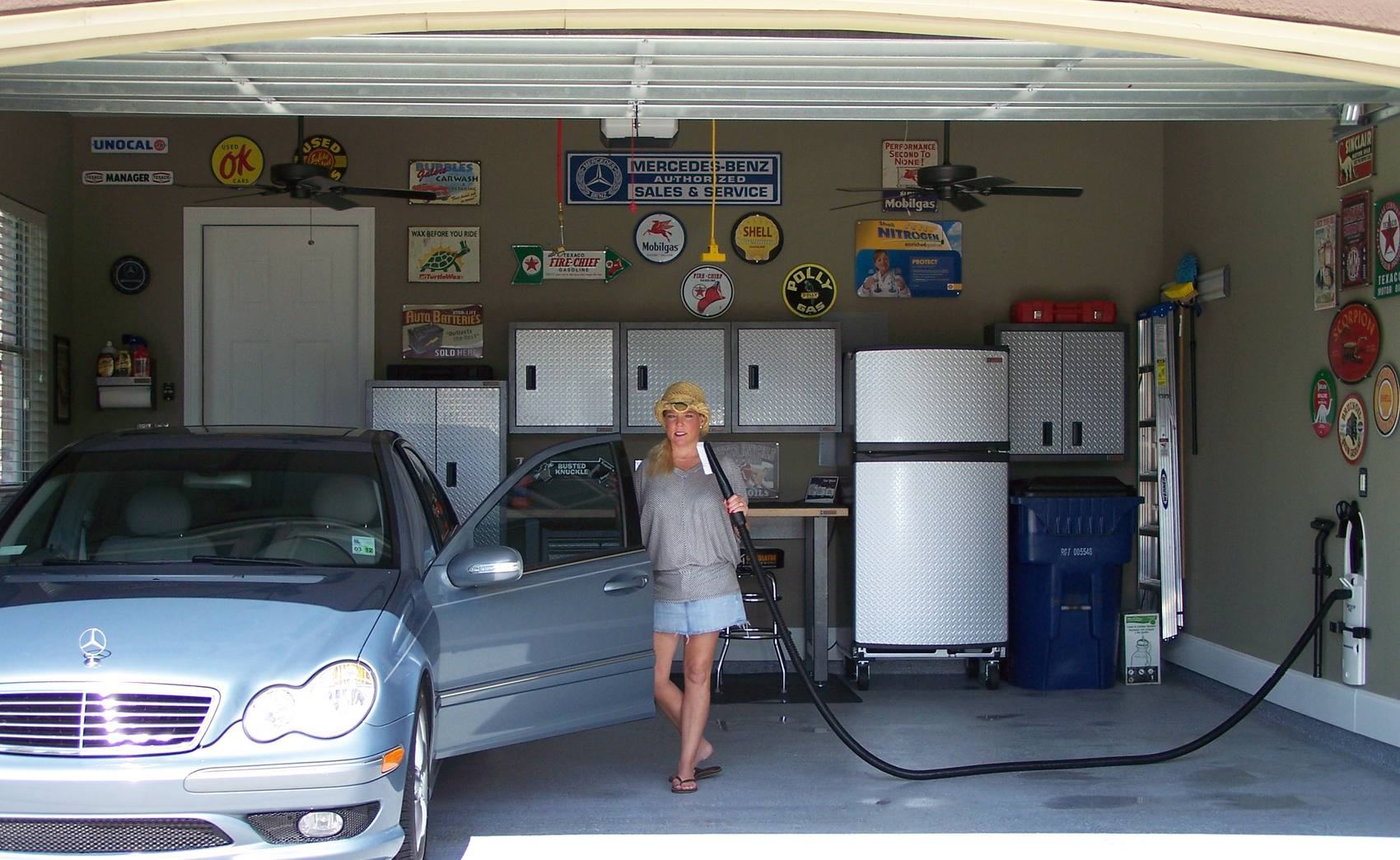A finished two-car garage with an epoxy floor and a wall of vintage motor-oil signs. A GarageVac is mounted on the right-hand wall, its hose stretched across to a Mercedes with the driver's door open.