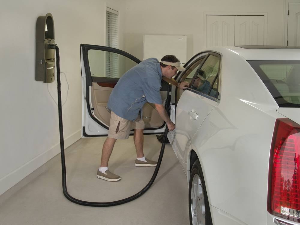 A GarageVac mounted on a garage wall with the hose run to the open driver's door of a white sedan being vacuumed.